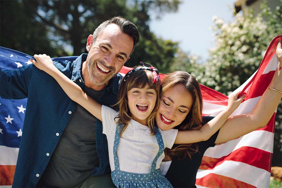 Personal Insurance - Closeup Portrait of a Cheerful Family with a Young Daughter Standing Outside in the Backyard While Holding an American Flag