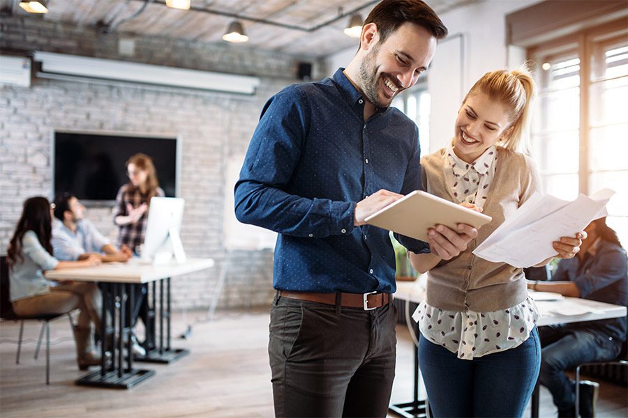 Commercial Insurance - Professional Showing another Something on their Tabelt and They are Both Smiling While Standing in the Office with Other Coworks While Light is Pouring In